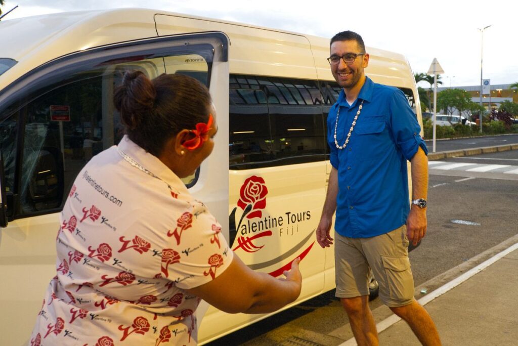 Explore Fiji's Must See Attractions With Local Tour Operators: All You Need To Know A friendly Valentine Tours Fiji staff member welcoming a customer during an airport transfer in Fiji. The staff member, smiling and extending a hand in greeting, stands next to a Valentine Tours Fiji vehicle, highlighting the company's airport transfers Fiji service.